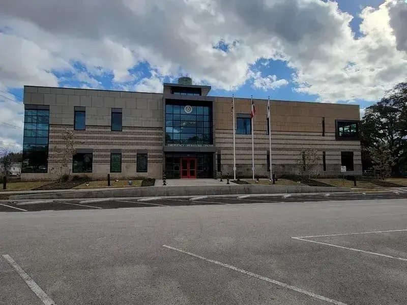 A tan and grey two-story brick office building with a central glass entryway and three flagpoles against a cloudy sky.