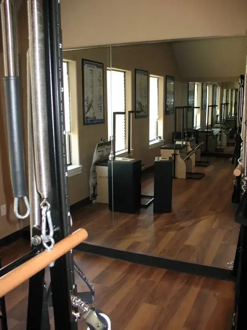 A brightly lit Pilates studio with wooden floors, reflecting equipment in a large mirror against a beige wall.