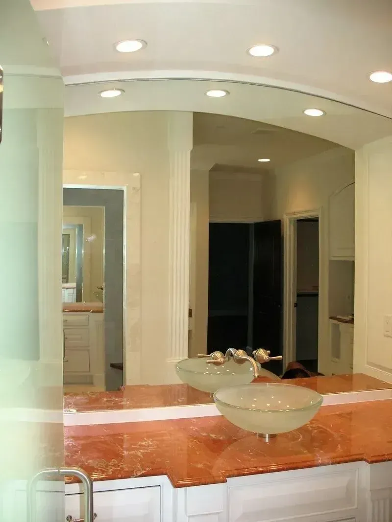 A bathroom vanity featuring a reddish-orange stone countertop, two glass vessel sinks, and a large, curved wall mirror.