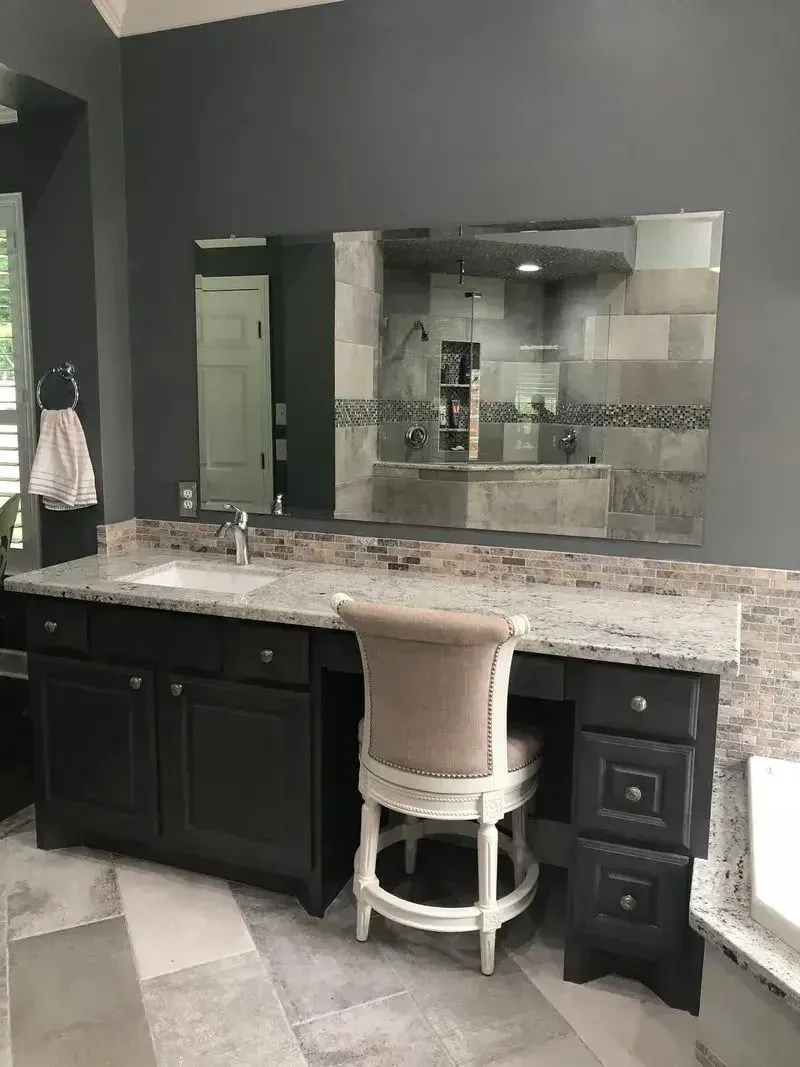 A dark grey bathroom vanity with a speckled granite countertop, a large wall mirror, and a light-colored stool.