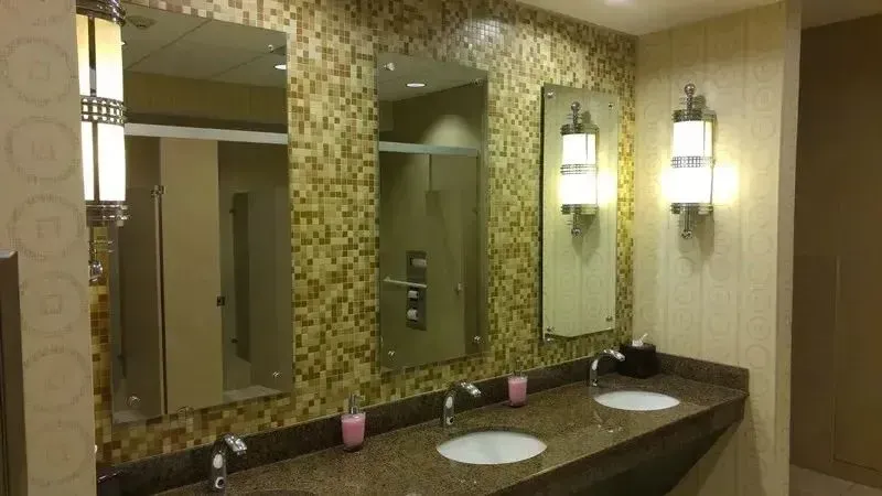 A clean public bathroom vanity with two white sinks, chrome faucets, three mirrors, and elegant wall sconces on gold tile.