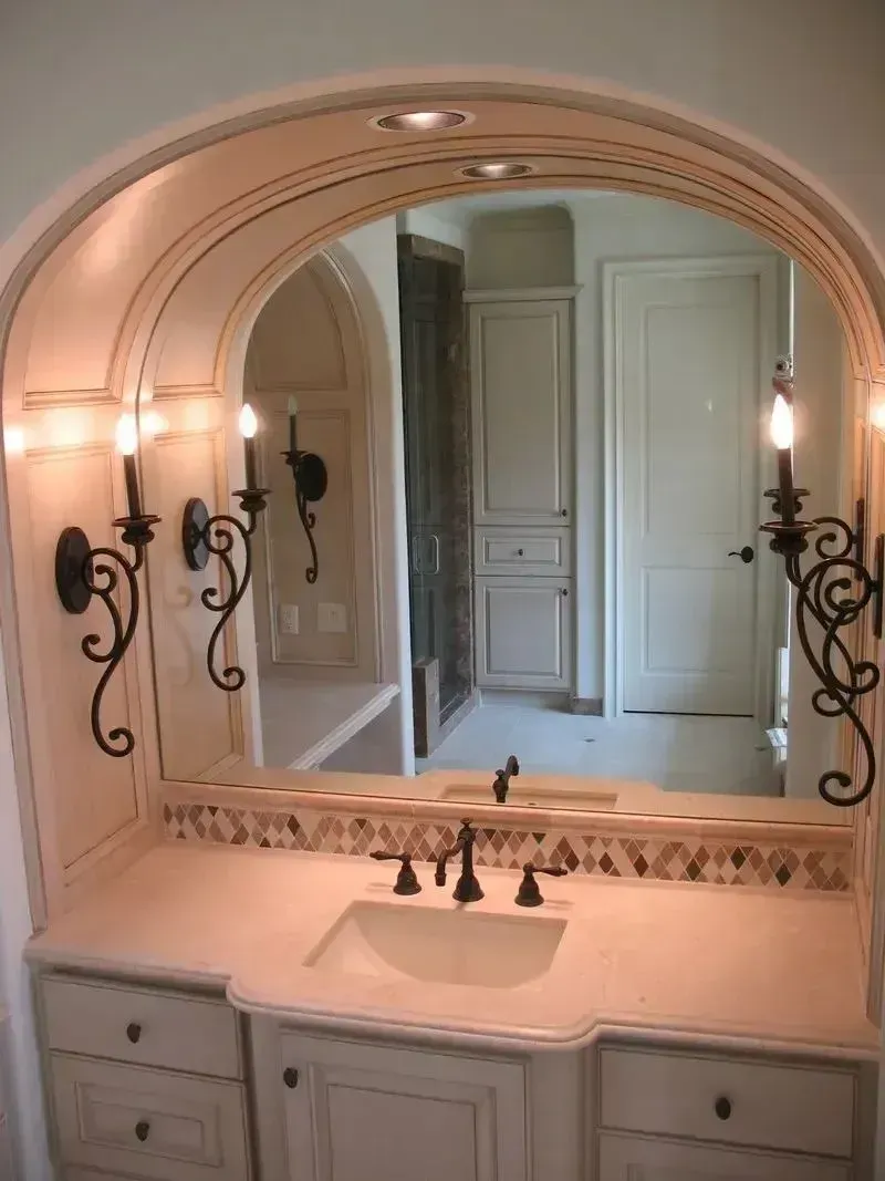 A bathroom vanity with an arched mirror, ornate iron wall sconces, a stone countertop, and light-colored cabinetry.