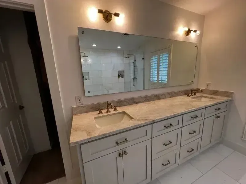 A double-sink bathroom vanity with light stone countertops, white cabinets, and a wide mirror under two wall sconces.