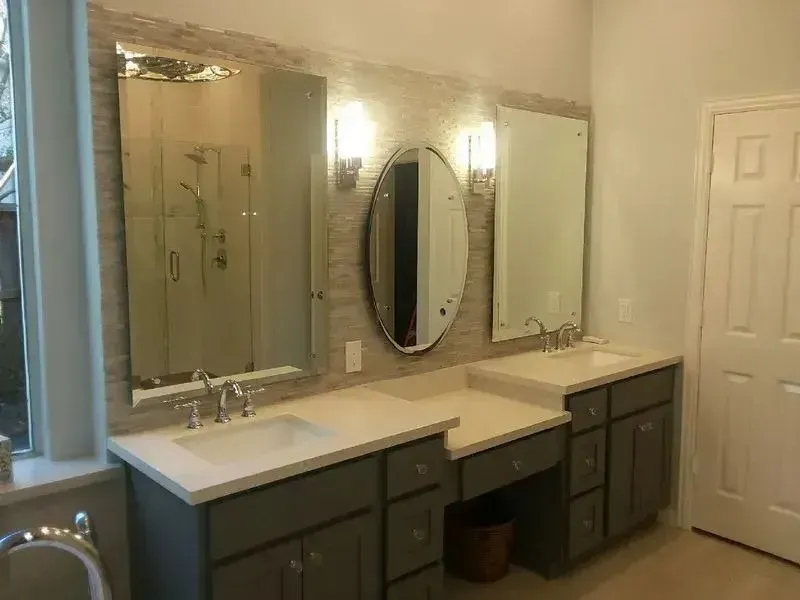 Bathroom vanity with gray cabinets, white countertop, three mirrors, and wall sconces on a textured stone-tiled wall.