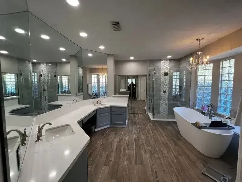 A modern, spacious bathroom with gray cabinetry, a white countertop, wood-look tile floors, and a freestanding tub.