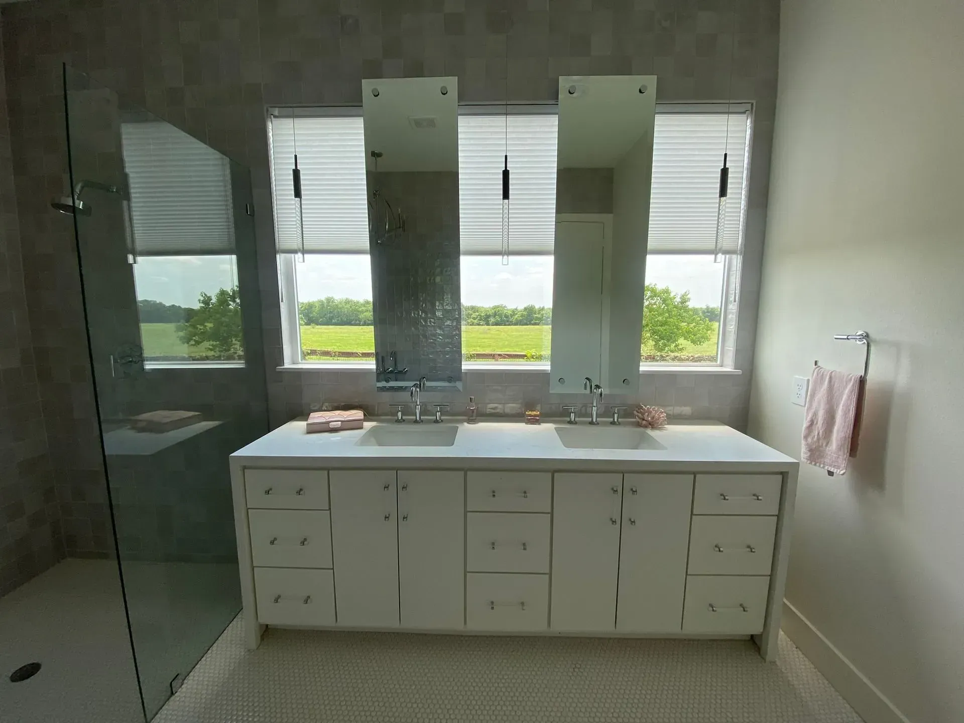A modern bathroom featuring a white double vanity, two vertical rectangular mirrors, large windows, and a glass shower.