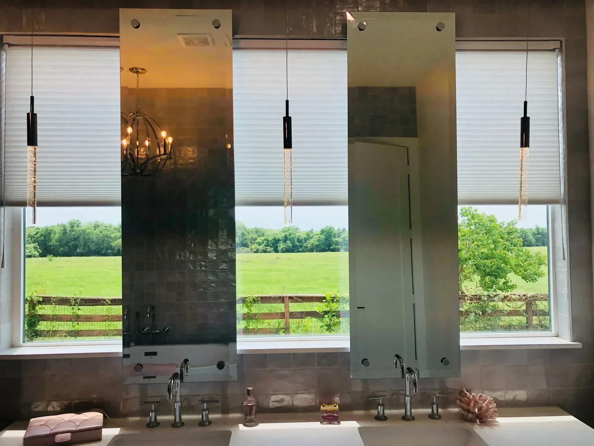 Two vertical rectangular mirrors hang above a double bathroom vanity in front of a wide window overlooking a field.