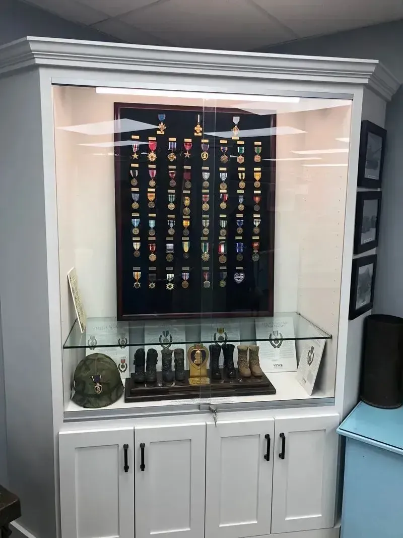 White display cabinet with glass shelves, medals, and military memorabilia under a framed display board.