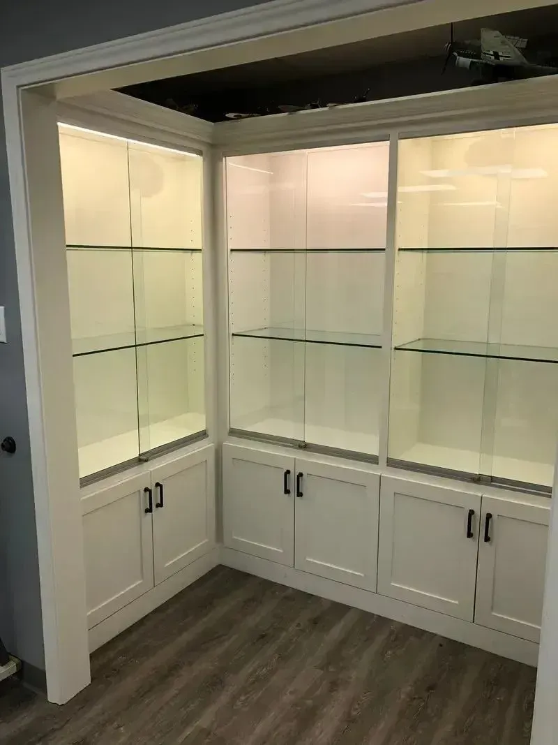 Built-in corner display cabinets with glass shelves, white lower cabinets, and wood flooring.