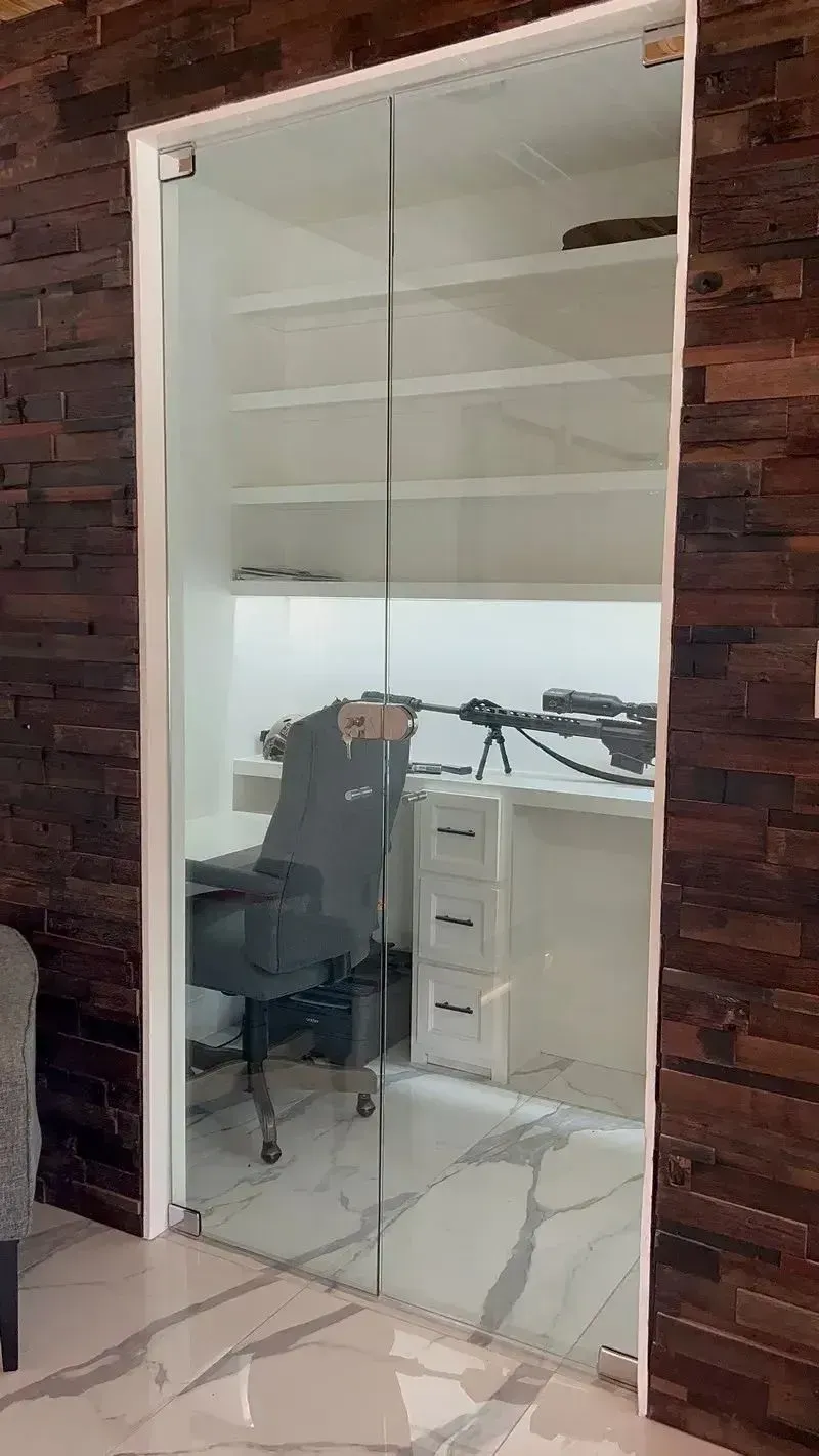 A frameless glass double door revealing a small office room with a desk, chair, shelves, and a rifle on the desk.