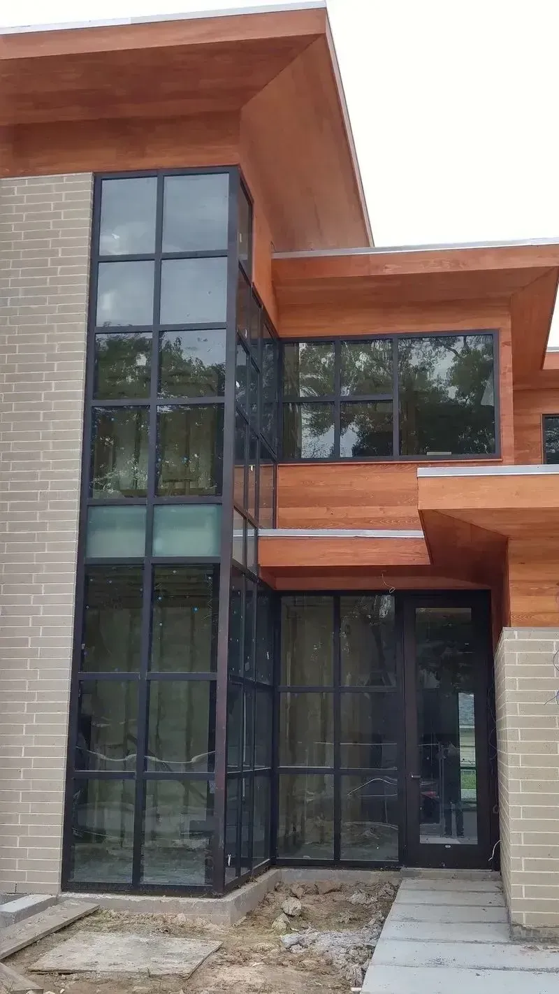 Modern home exterior featuring a floor-to-ceiling glass corner window, tan brick walls, and cedar wood siding.