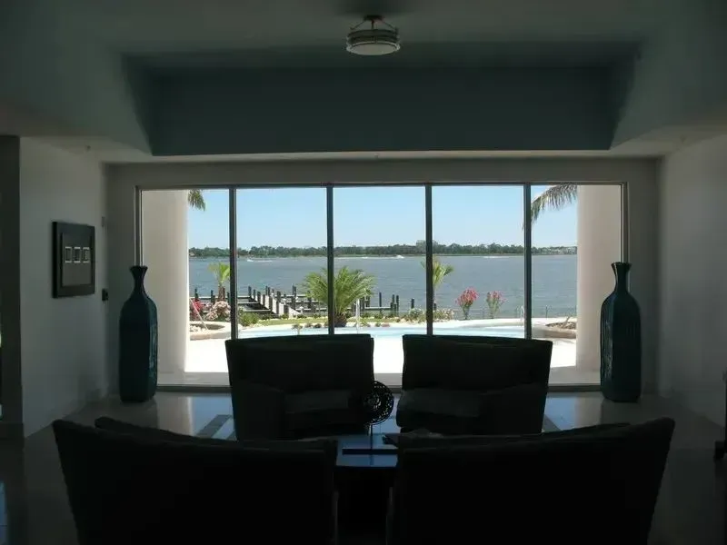 A living room with dark seating faces a large window overlooking a swimming pool, a dock, and a wide body of water.