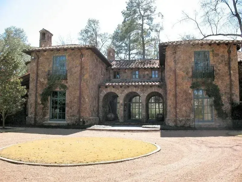 A two-story stone mansion with a Mediterranean-style roof and arched entryways, set behind a circular gravel driveway.