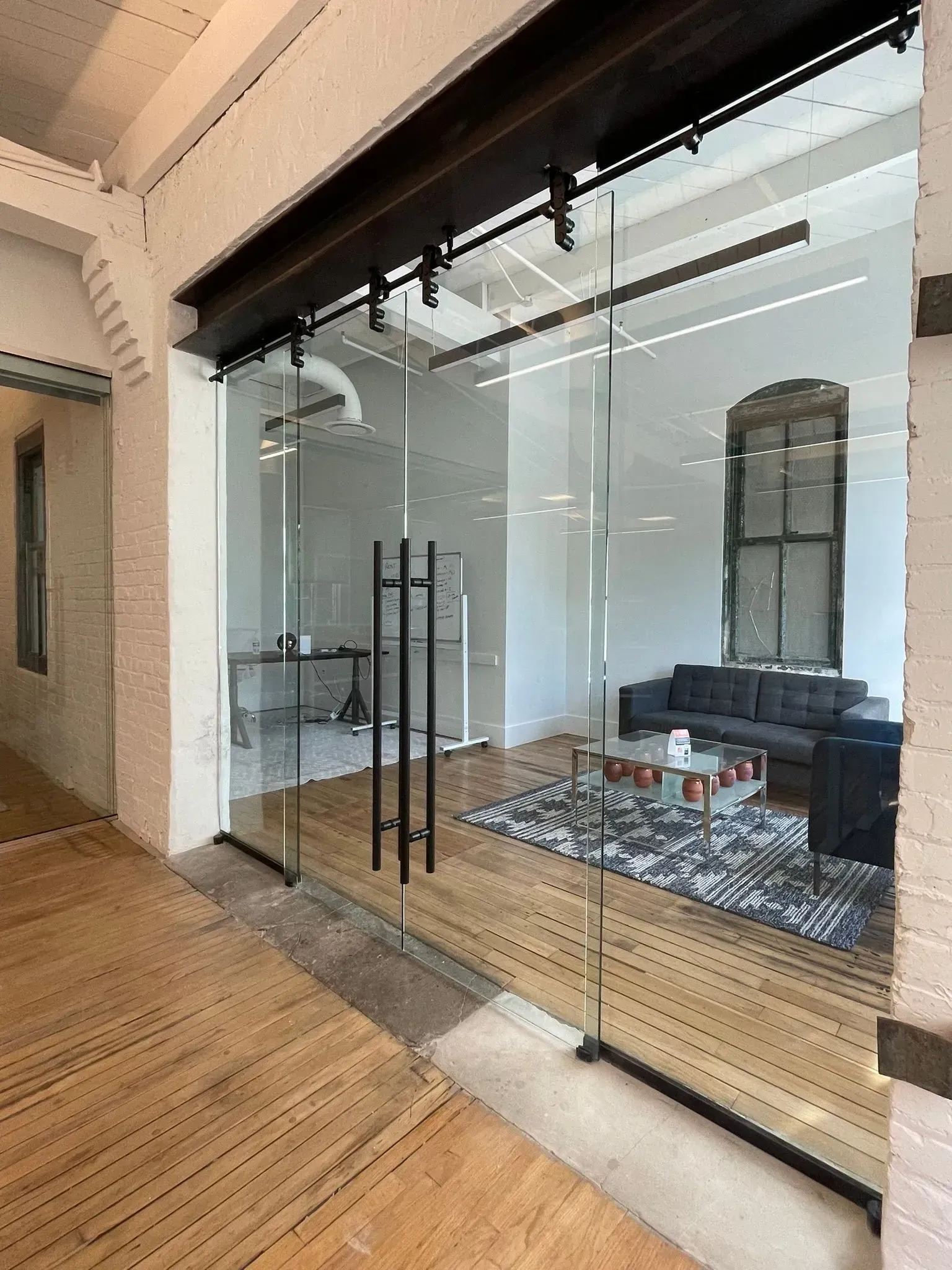 A sliding glass office door with black hardware, set in a room with white brick walls, hardwood floors, and a sofa.