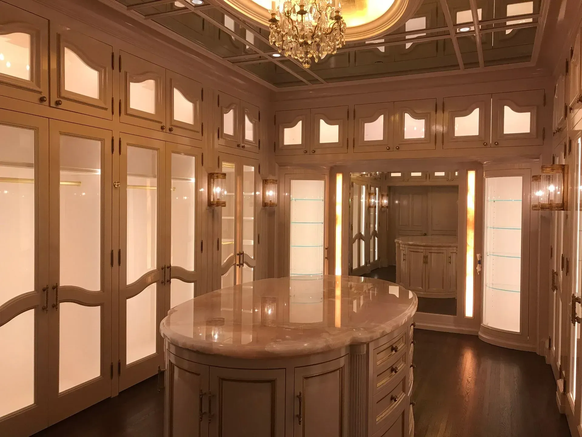 A luxurious, well-lit walk-in closet with cream-colored cabinetry, frosted glass doors, and a central marble-top island.
