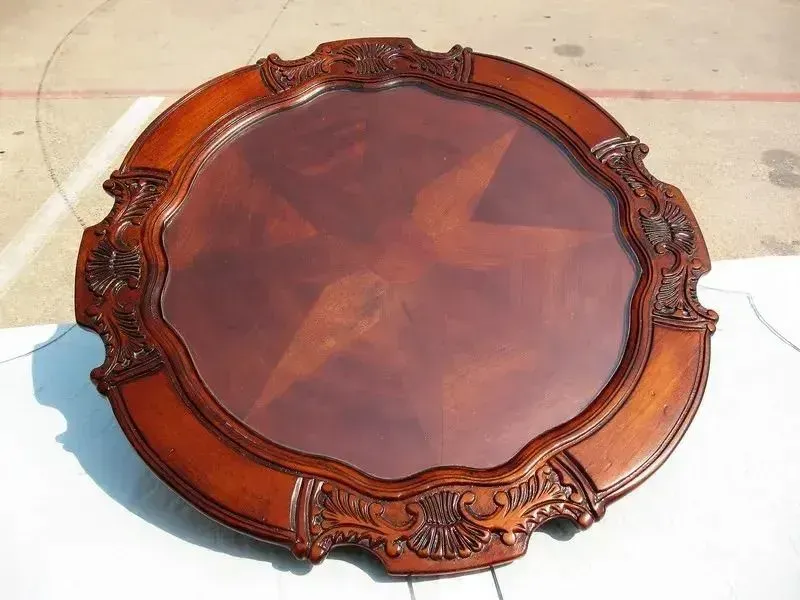 A round, wooden table top with ornate, carved decorative edges and a starburst wood-veneer center pattern.