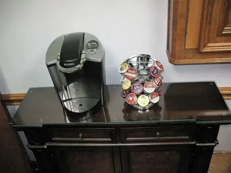 A Keurig coffee maker and a circular pod holder filled with various coffee capsules sit on a dark wooden cabinet.
