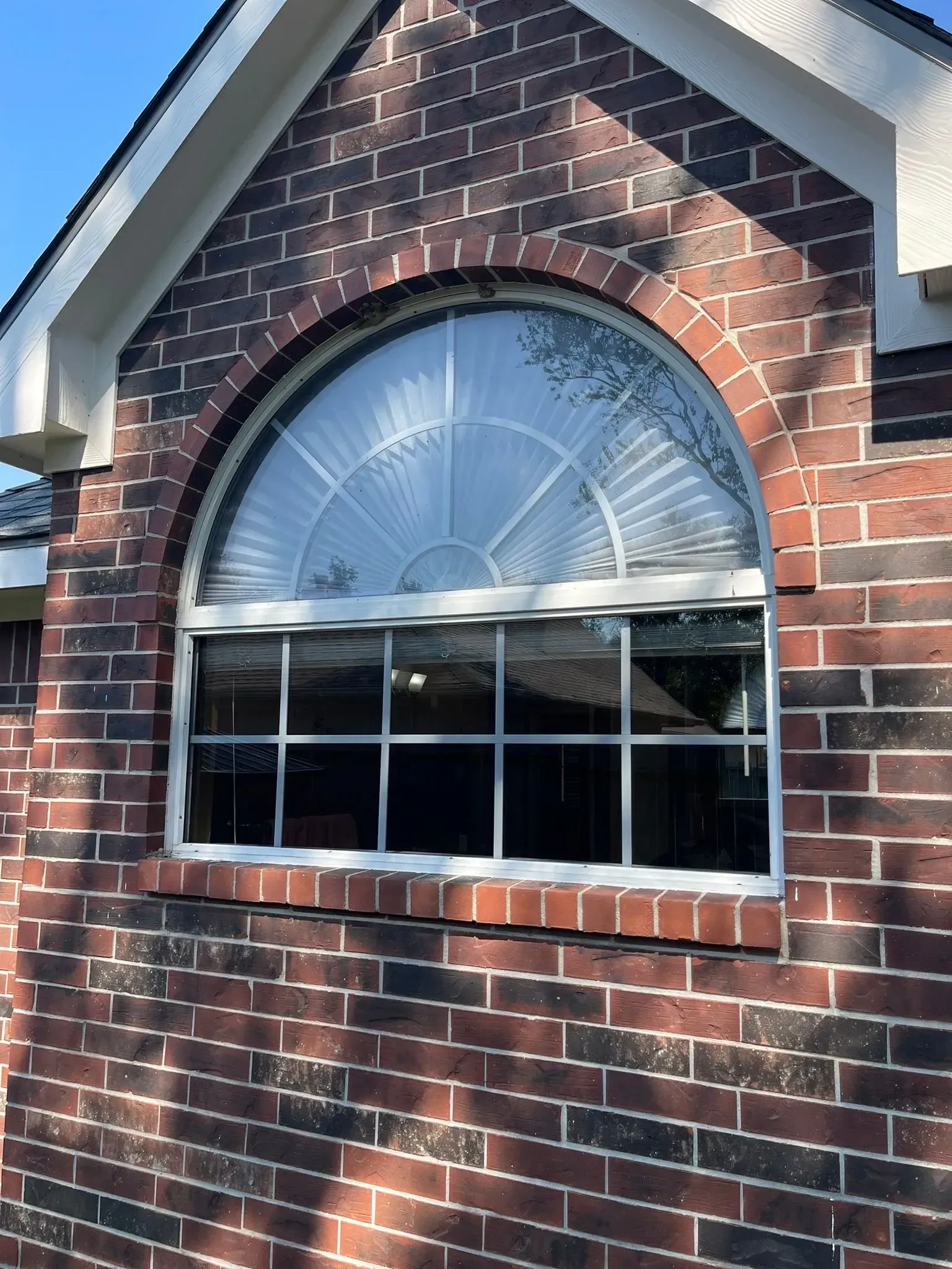 An arched window with a fan-shaped top pane and a grid-patterned lower section, set in a dark red brick wall.