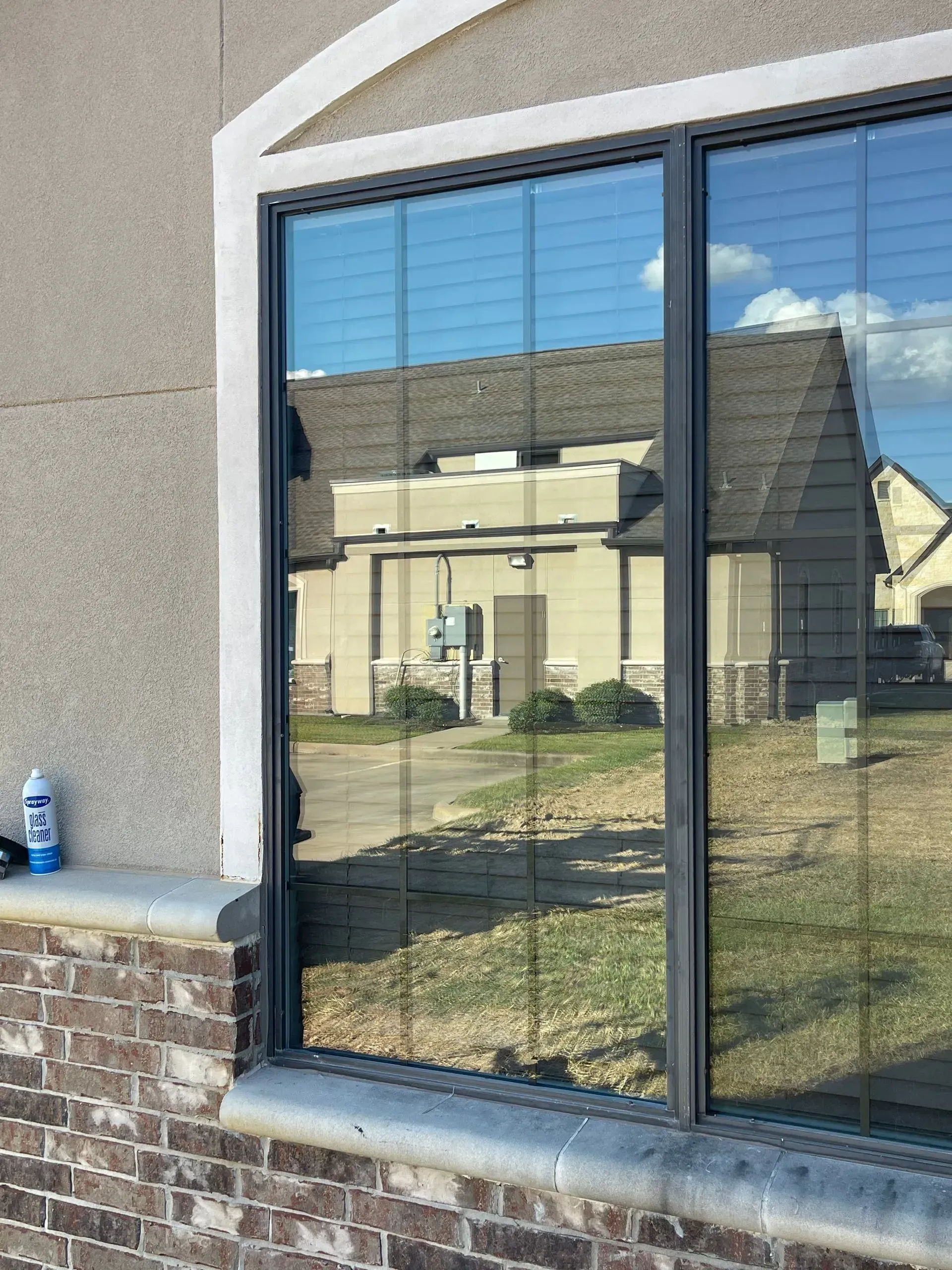 A window in a tan stucco wall with a brick base reflects a neighboring house and blue sky.