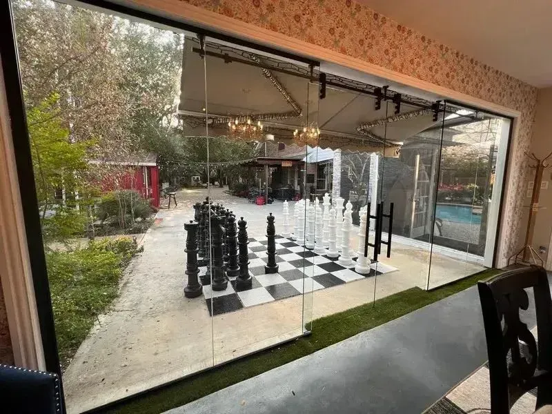 A large outdoor chessboard with giant black and white pieces is set on a patio, viewed through a glass sliding door.