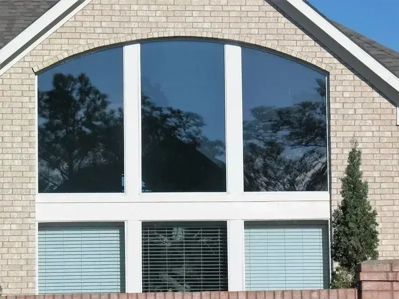A large, arched window with white frames set into a beige brick exterior wall, with reflections visible in the glass.