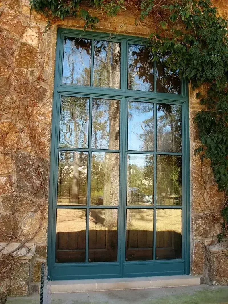 A tall, dark teal, multi-pane window with a decorative border set into a rustic, textured stone wall with ivy.
