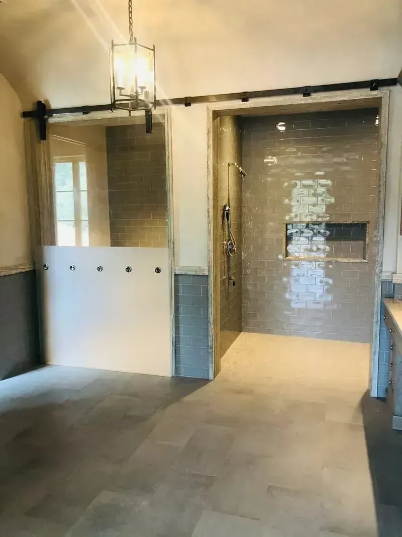 A bathroom features two shower stalls with sliding glass doors, neutral floor tiles, a wall-mounted shower head, and lamps.