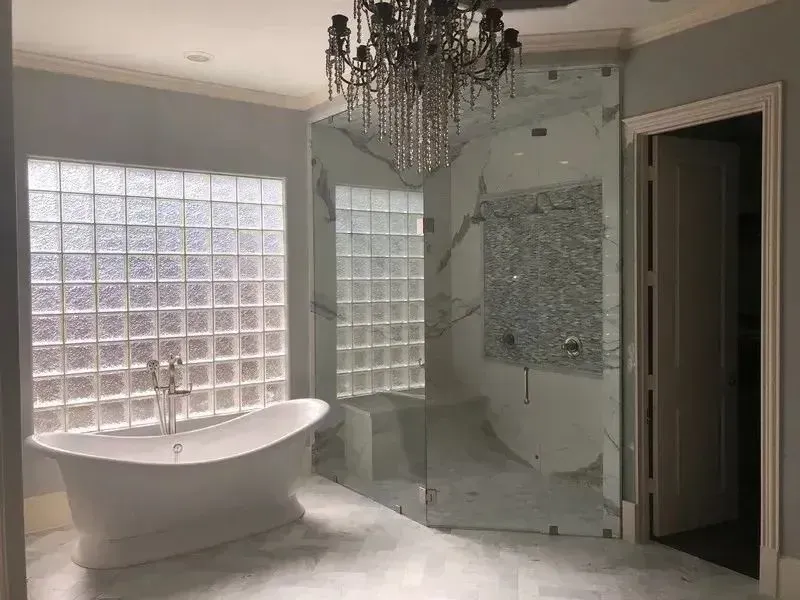 A modern bathroom featuring a white freestanding tub, a glass-enclosed shower with marble walls, and a crystal chandelier.