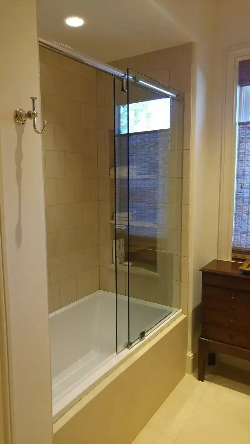 A bathroom featuring a white bathtub with a glass door, beige tiled walls, and a wooden vanity beside a window.