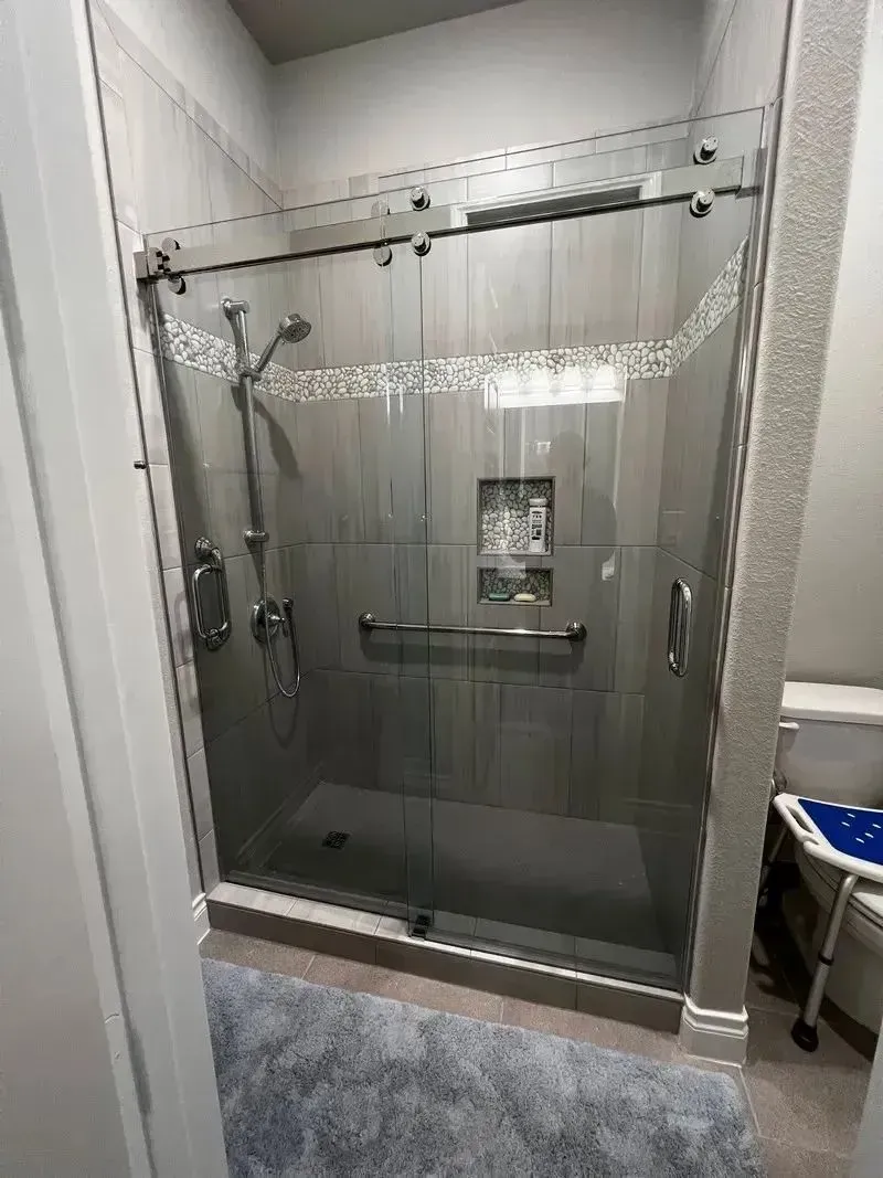 A walk-in shower with a glass sliding door, gray tiled walls, a handheld showerhead, built-in shelving, and a grab bar.