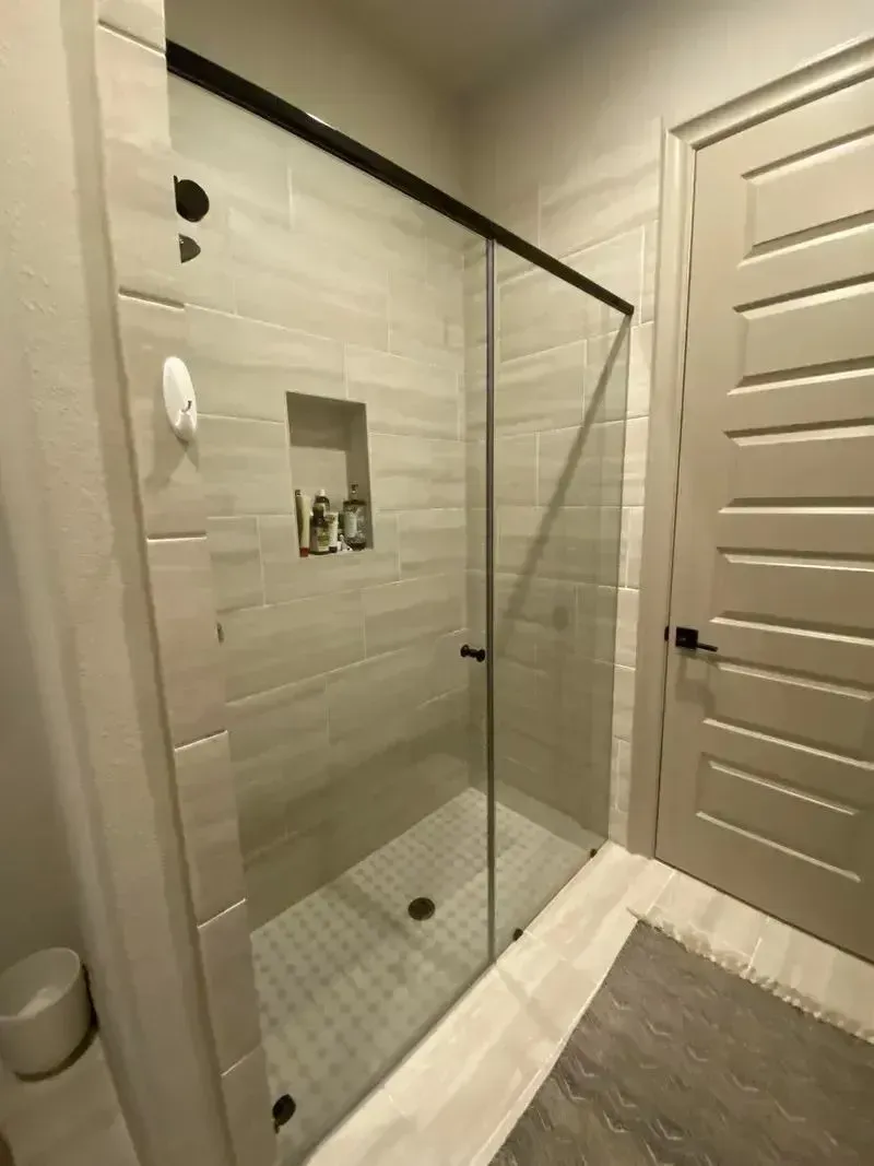 A modern walk-in shower with light-colored tile walls, a built-in shelf, glass door, and adjacent light-colored door.