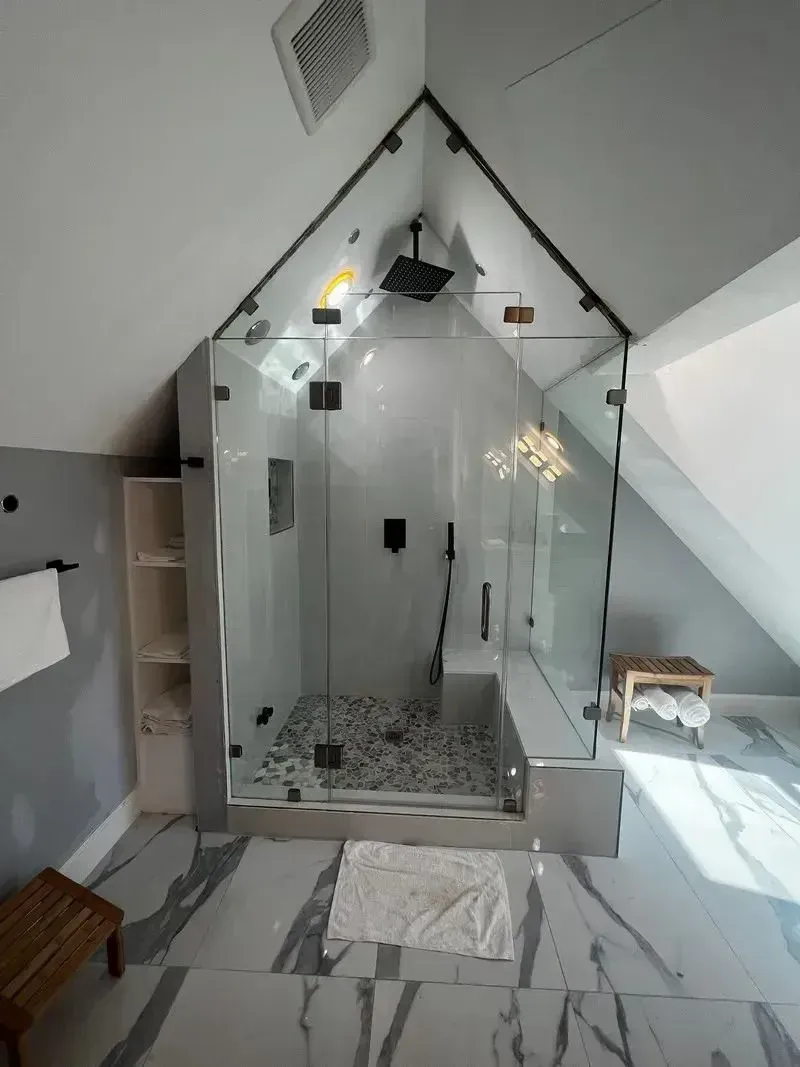 A modern glass-enclosed shower with pebble floor tiles, situated under a peaked roof in a room with marbled flooring.