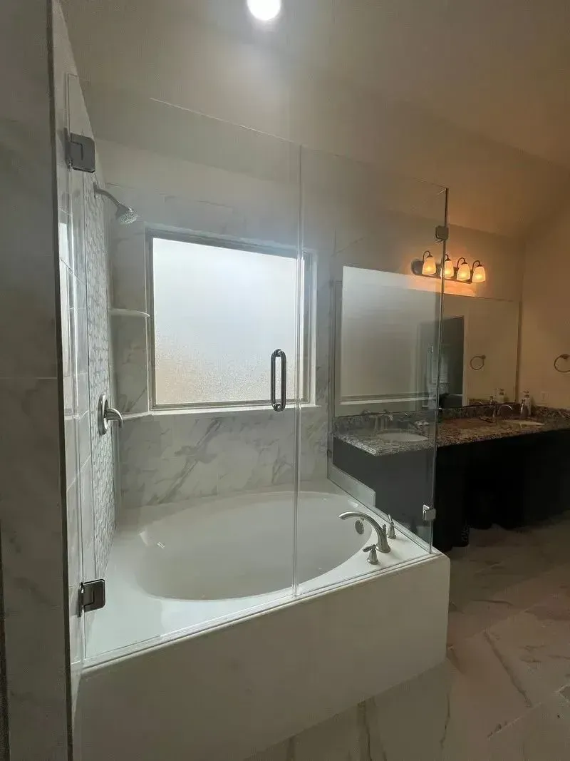 A modern bathroom featuring a white soaking tub with marble surround and glass shower doors in front of a frosted window.