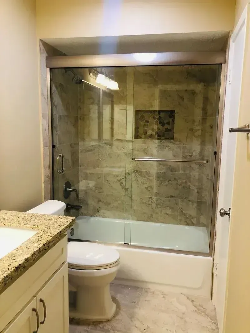A modern bathroom featuring a white toilet, a granite-topped vanity, and a bathtub with a glass sliding door.