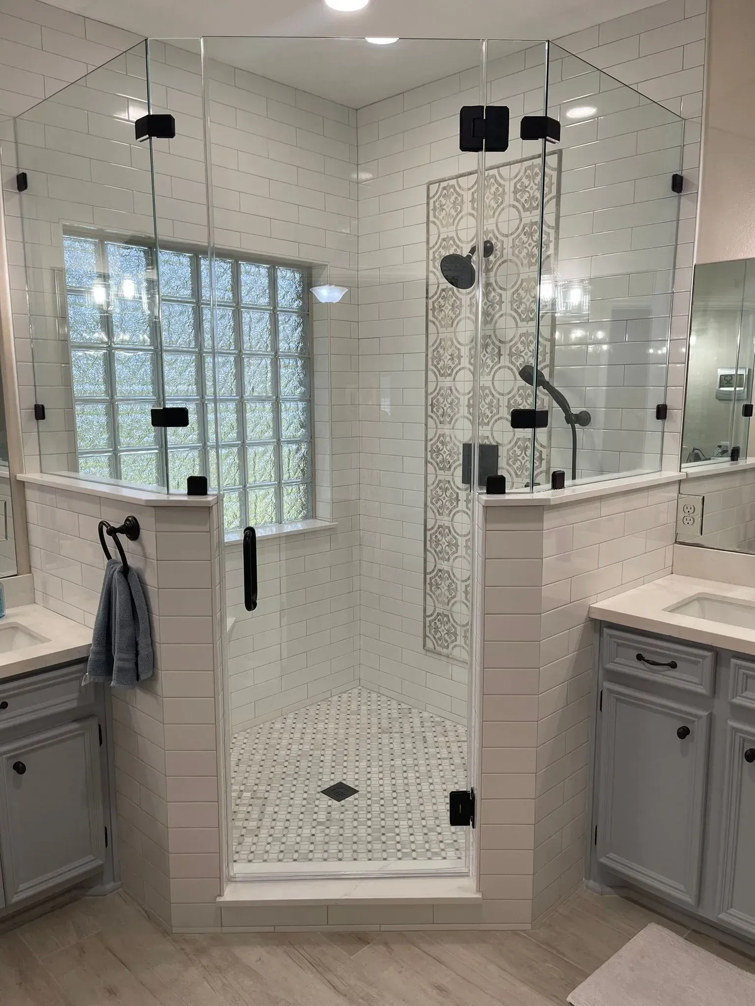 A glass-enclosed shower with white subway tile and a decorative patterned backsplash, situated between two gray vanities.