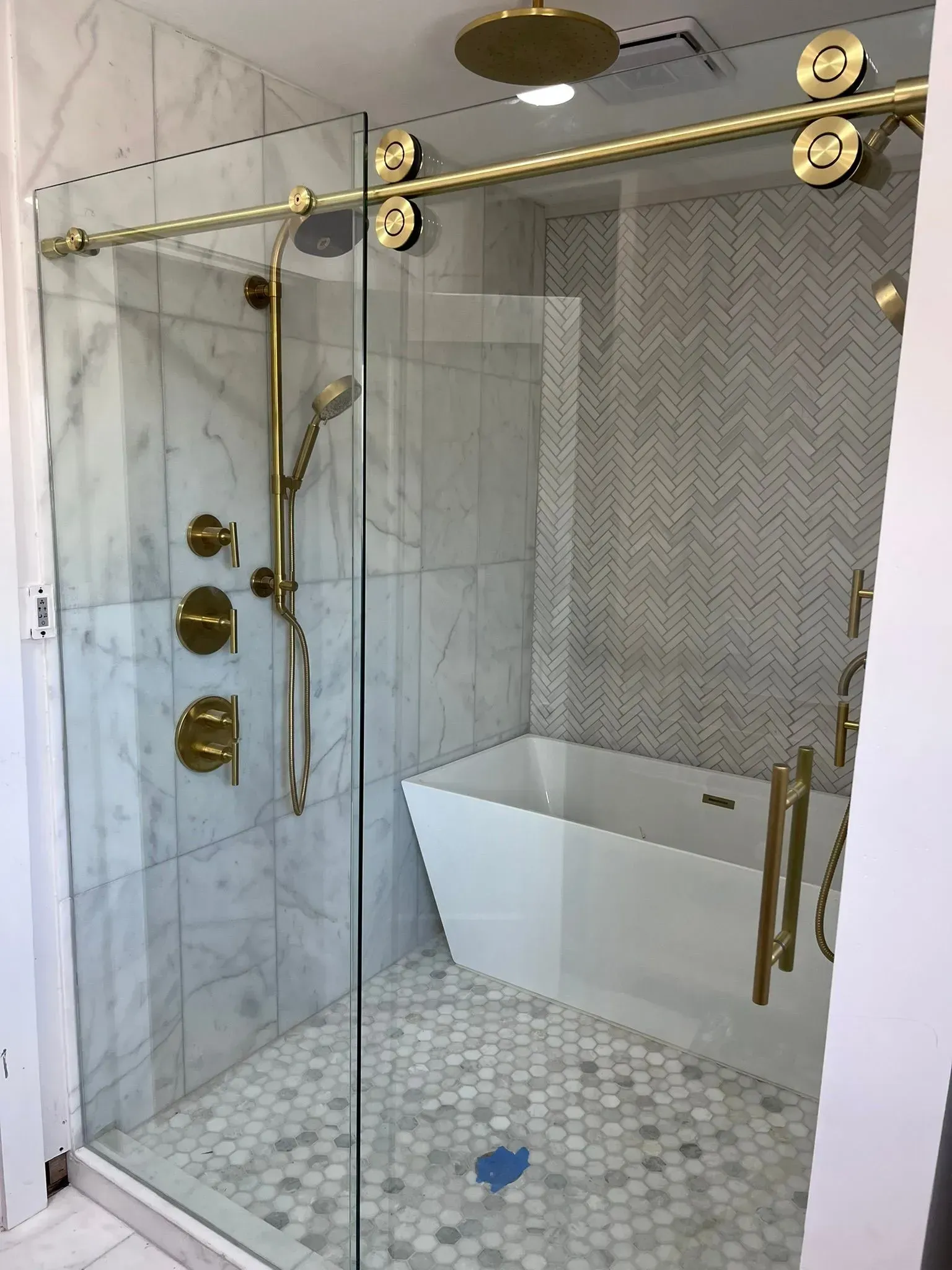 A modern walk-in shower with gold hardware, a freestanding bathtub, marble walls, and a herringbone tile accent wall.