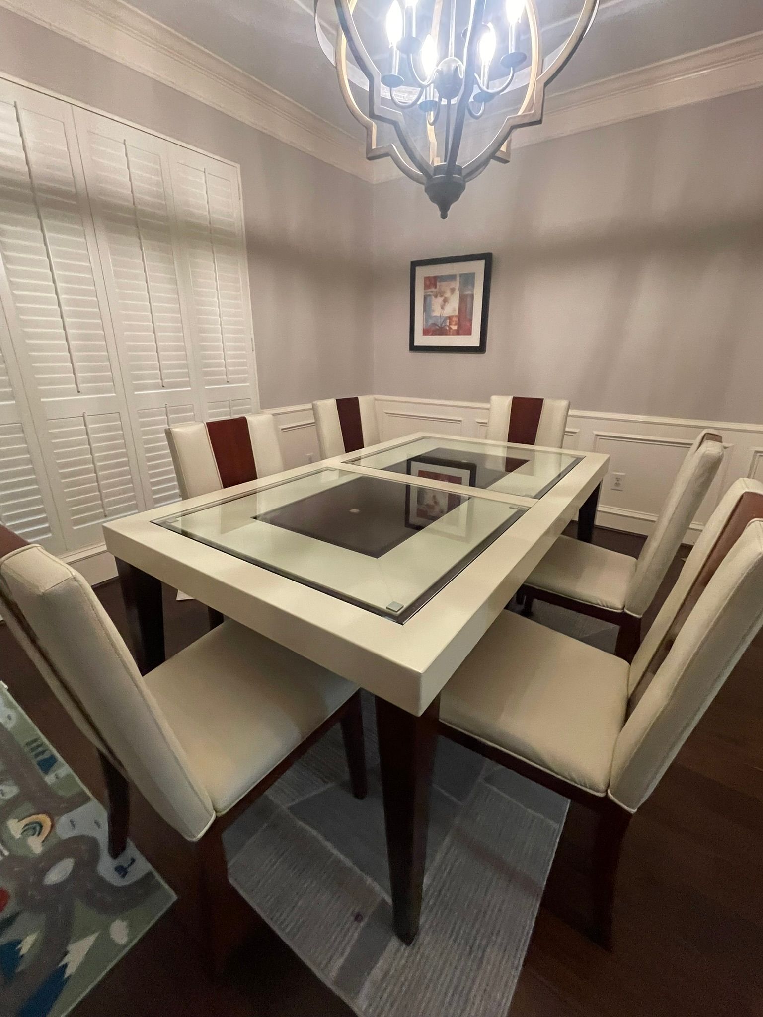 Dining room with white chairs around a glass-top table, chandelier overhead, and window blinds on the left.
