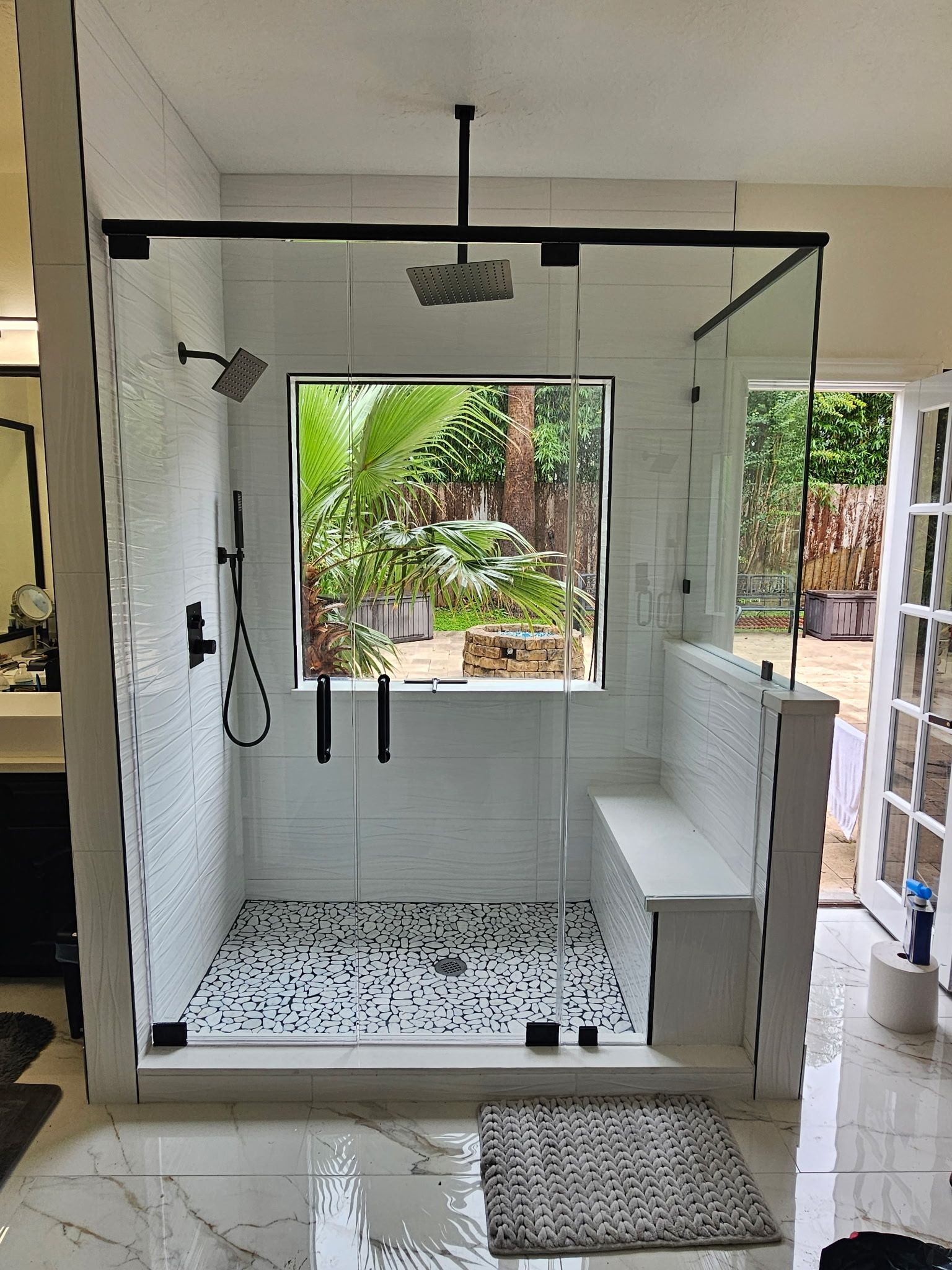 Modern outdoor shower stall with glass door, white walls, black fixtures, and pebble floor.