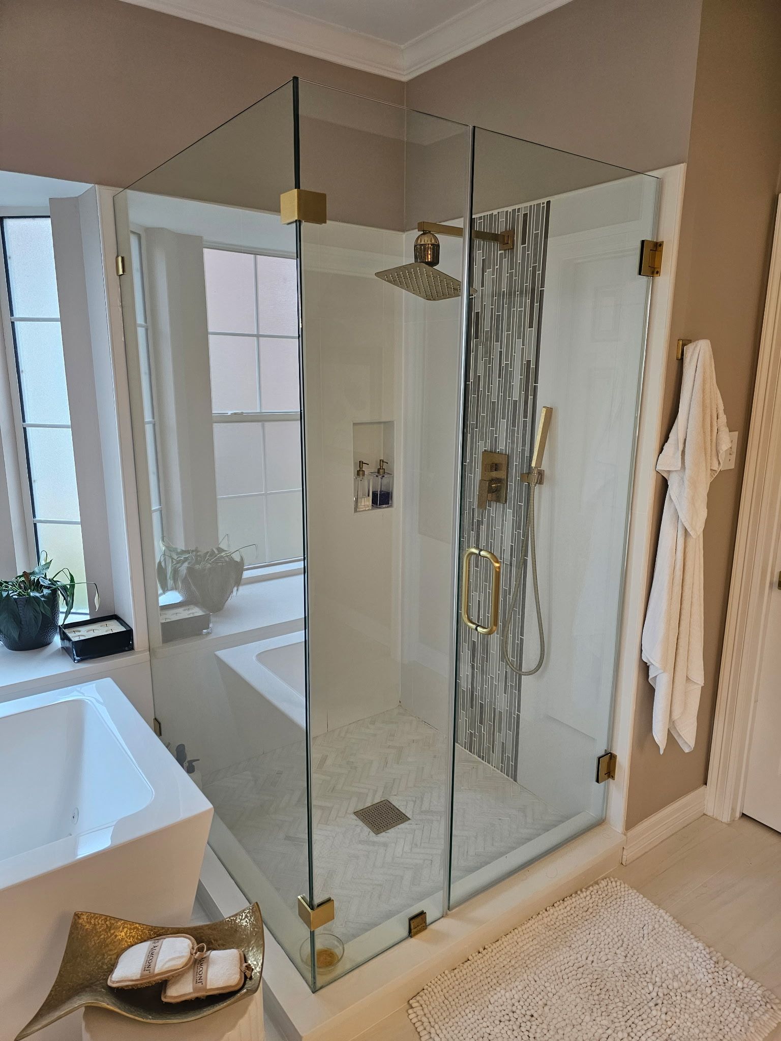 Glass shower enclosure in a bright bathroom with white tub, beige walls, and brass fixtures