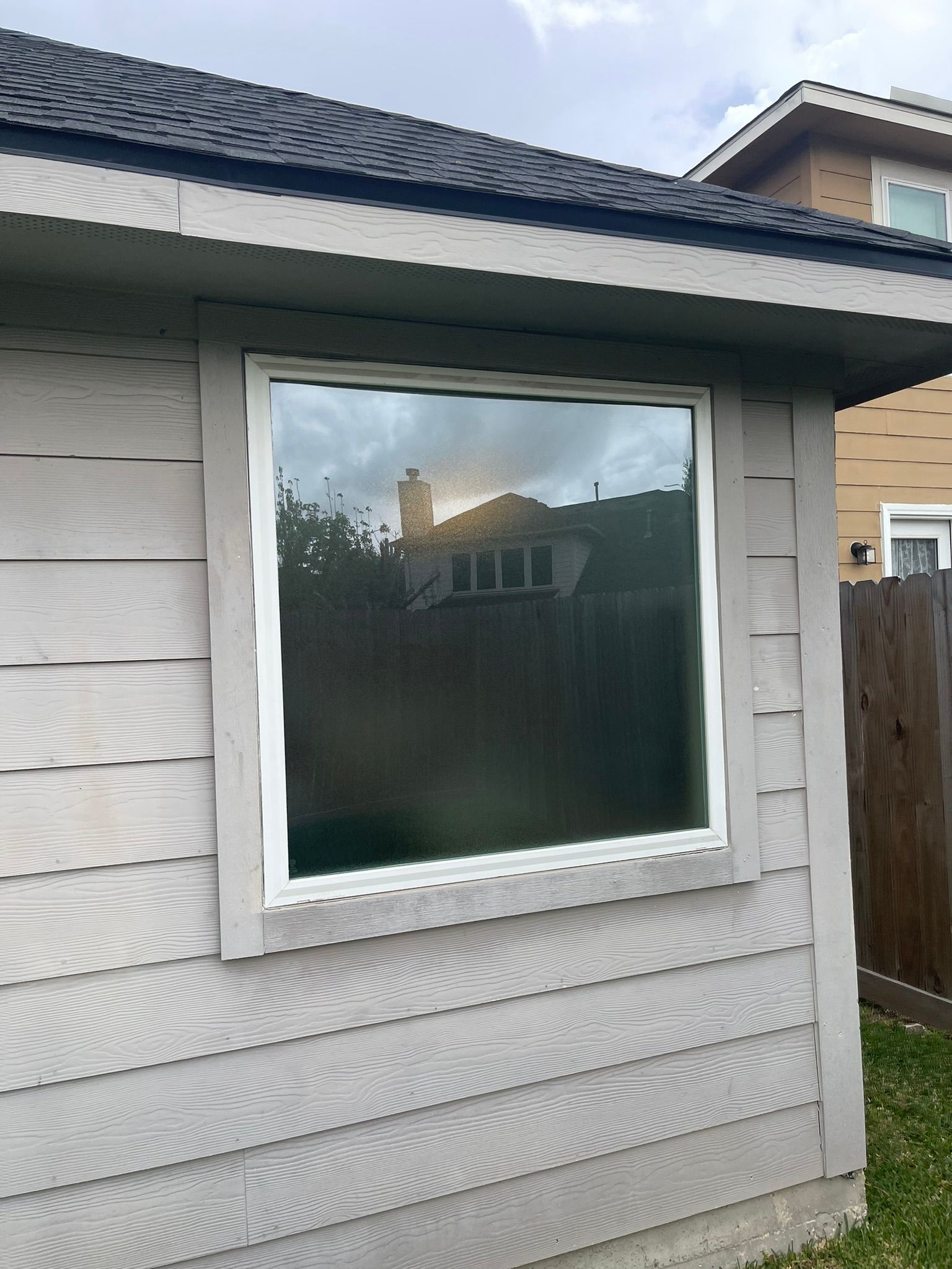 White-trimmed window on a gray-sided house, reflecting trees and a neighboring home.