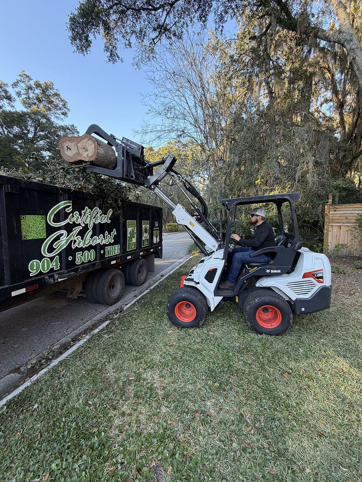 Skid steer loading debris into a black landscape trailer under trees on a grassy yard.