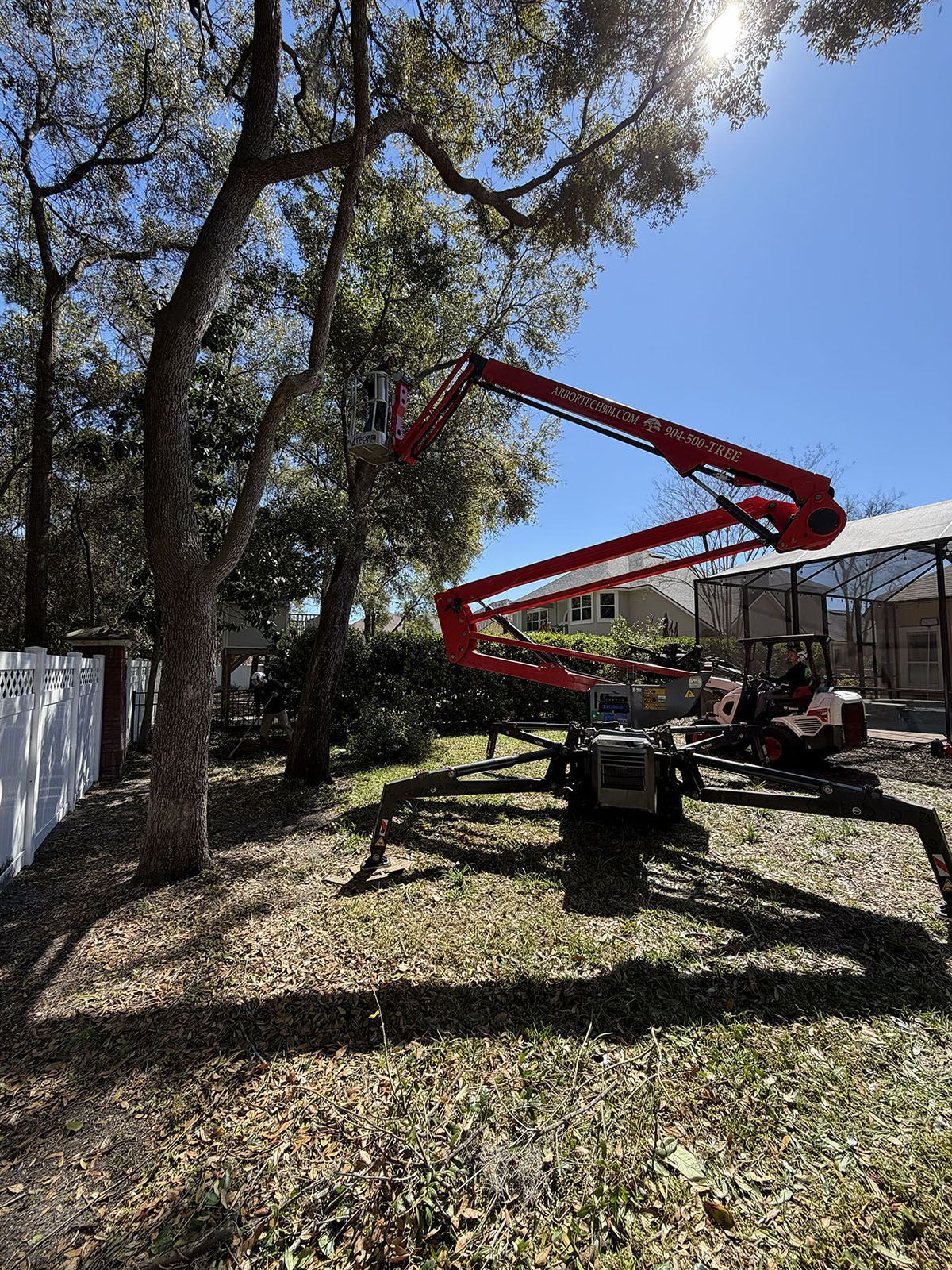 Red boom lift in a leafy yard beside a white fence and house, with a small utility vehicle nearby.