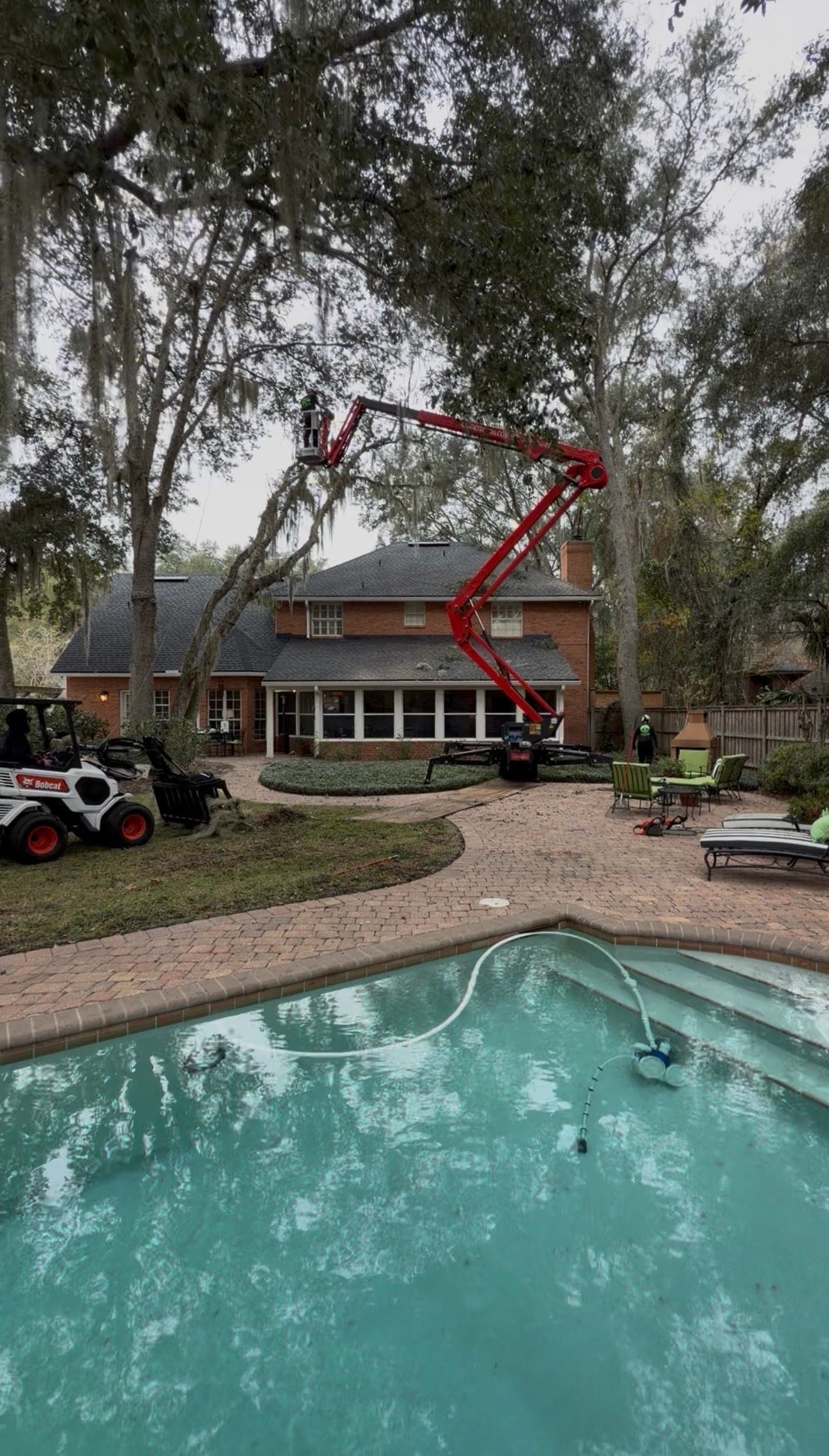 Backyard pool with a red boom lift pruning tall trees beside a house.