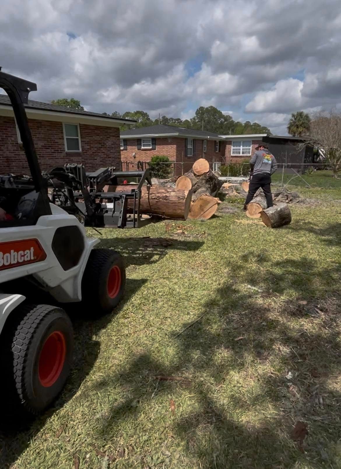 Worker with a Bobcat loader moving large logs in a yard beside a brick house under cloudy skies.