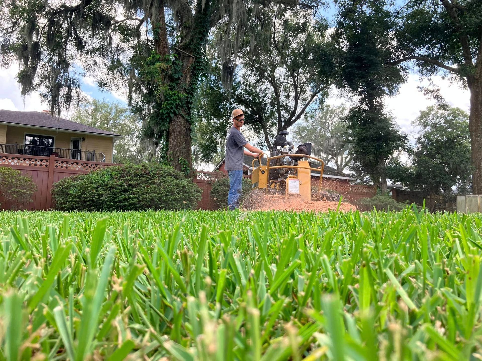 Person mowing grass in a yard with trees and a brick house in the background.