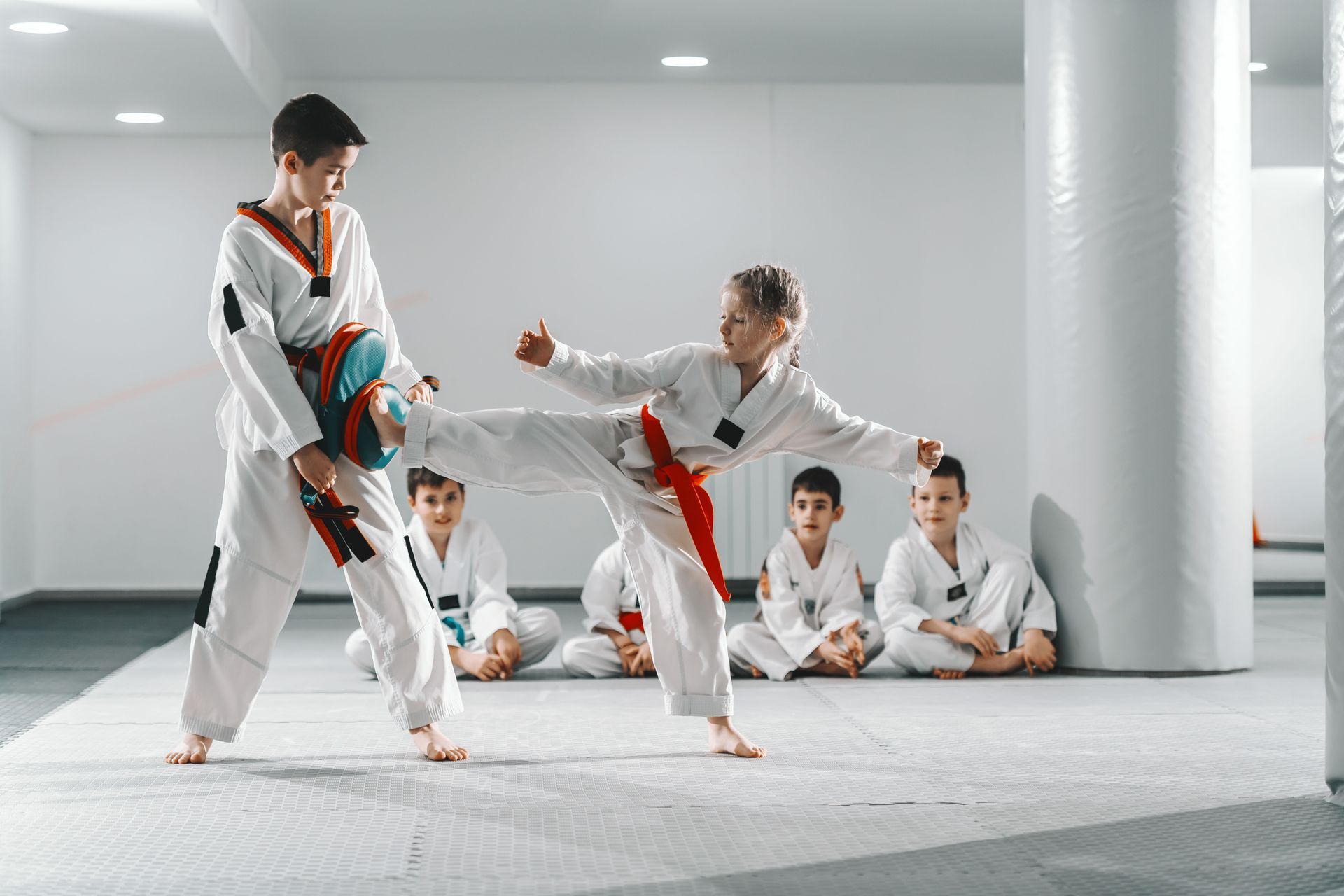 Two children in taekwondo uniforms practice kicking, others observe.
