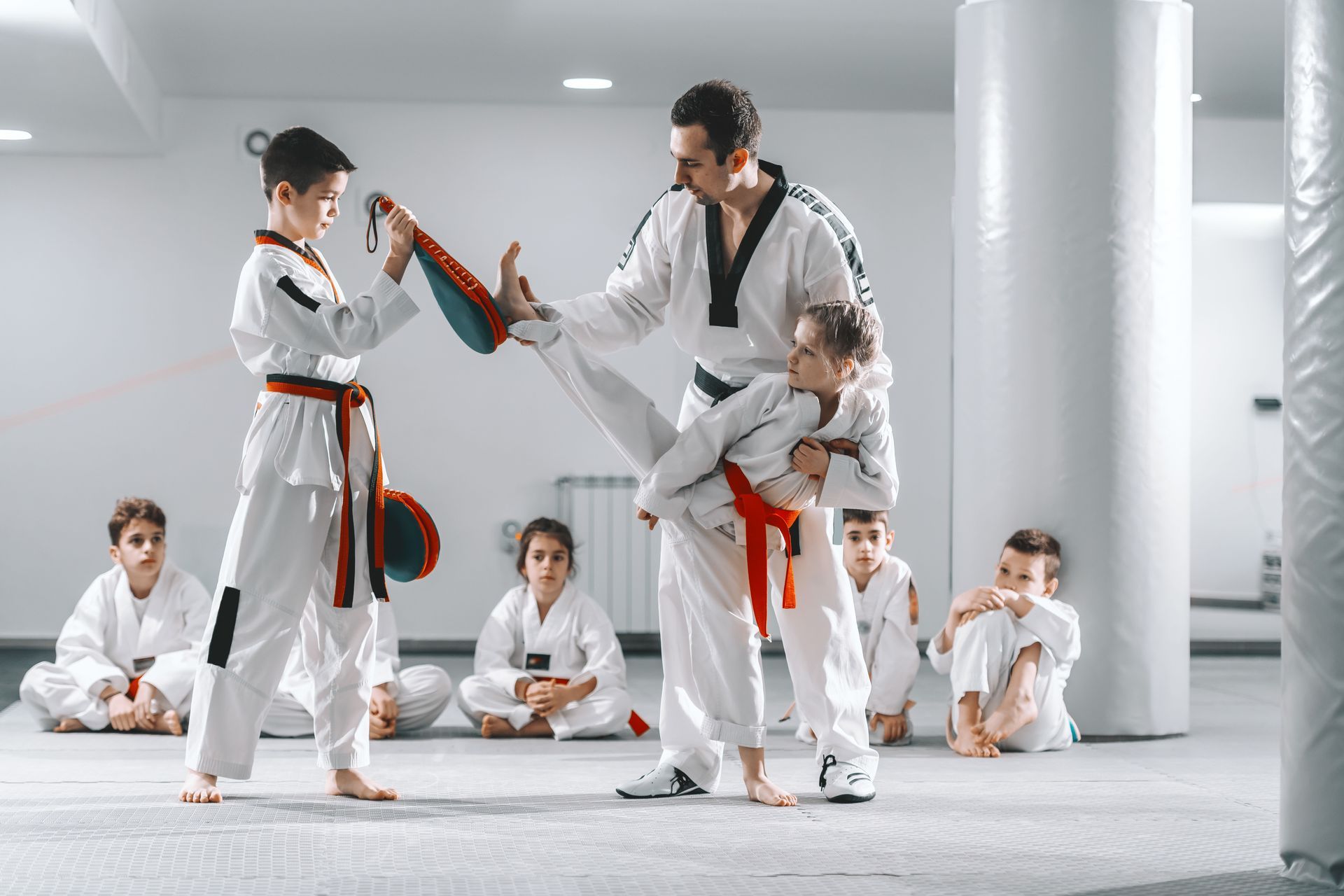 Martial arts students in white uniforms practice a kick. Instructor and others observe.