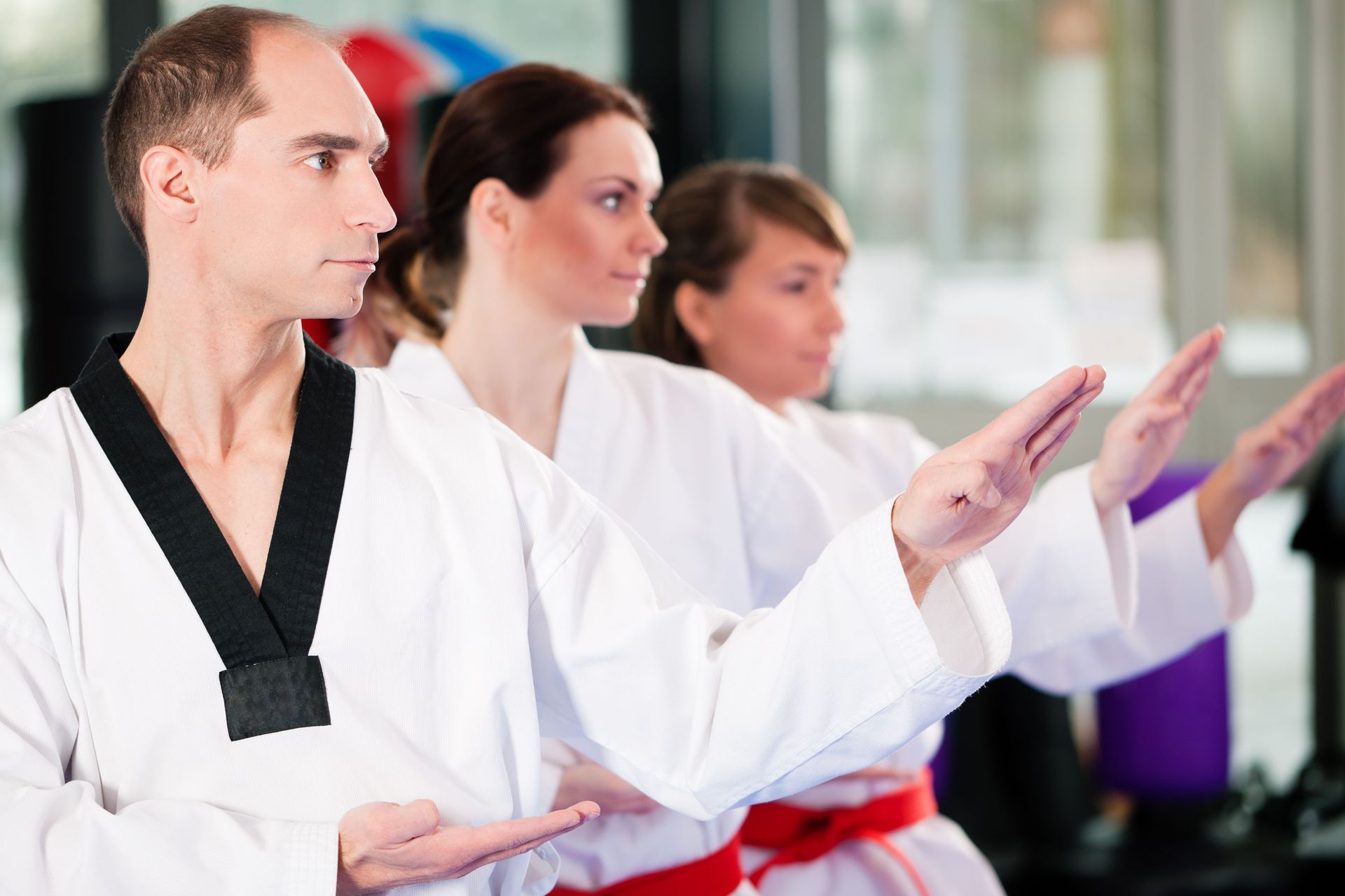 Three martial artists in white uniforms with black and red belts perform a synchronized hand strike in a studio.