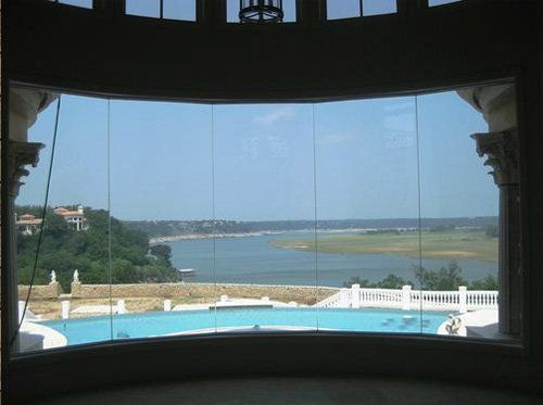 A panoramic view of a turquoise swimming pool overlooking a bay, framed by large, floor-to-ceiling glass windows.