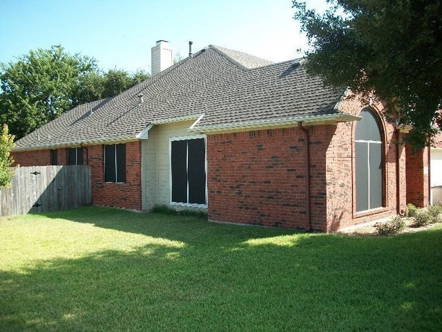 A single-story brick house with a gray shingle roof, dark window screens, and a green lawn under a clear blue sky.